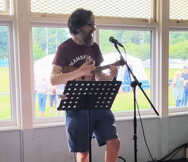 Eric playing a uke, on stage with microphone, wearing Mansfield Uke Group t-shirt