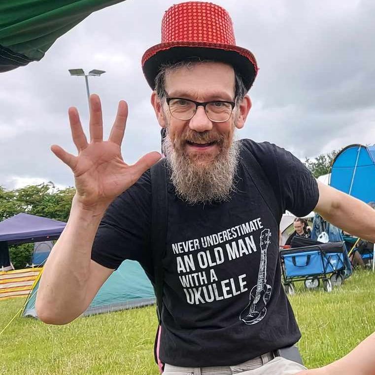 Eric, wearing red sparkly hat, in campsite, holding up his hand, with T shirt with the logo 'Never underestimate an old man with a ukulele'