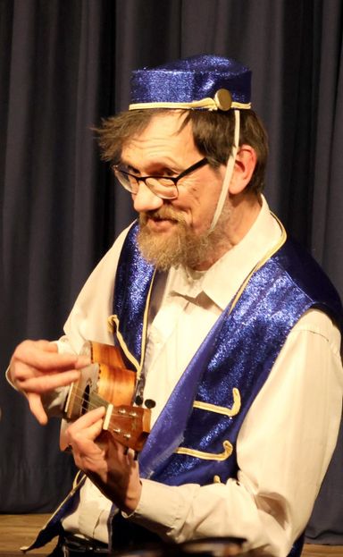 Side-profile picture of Eric playing uke in pantomime outfit, including small blue 'bell-boy' hat.