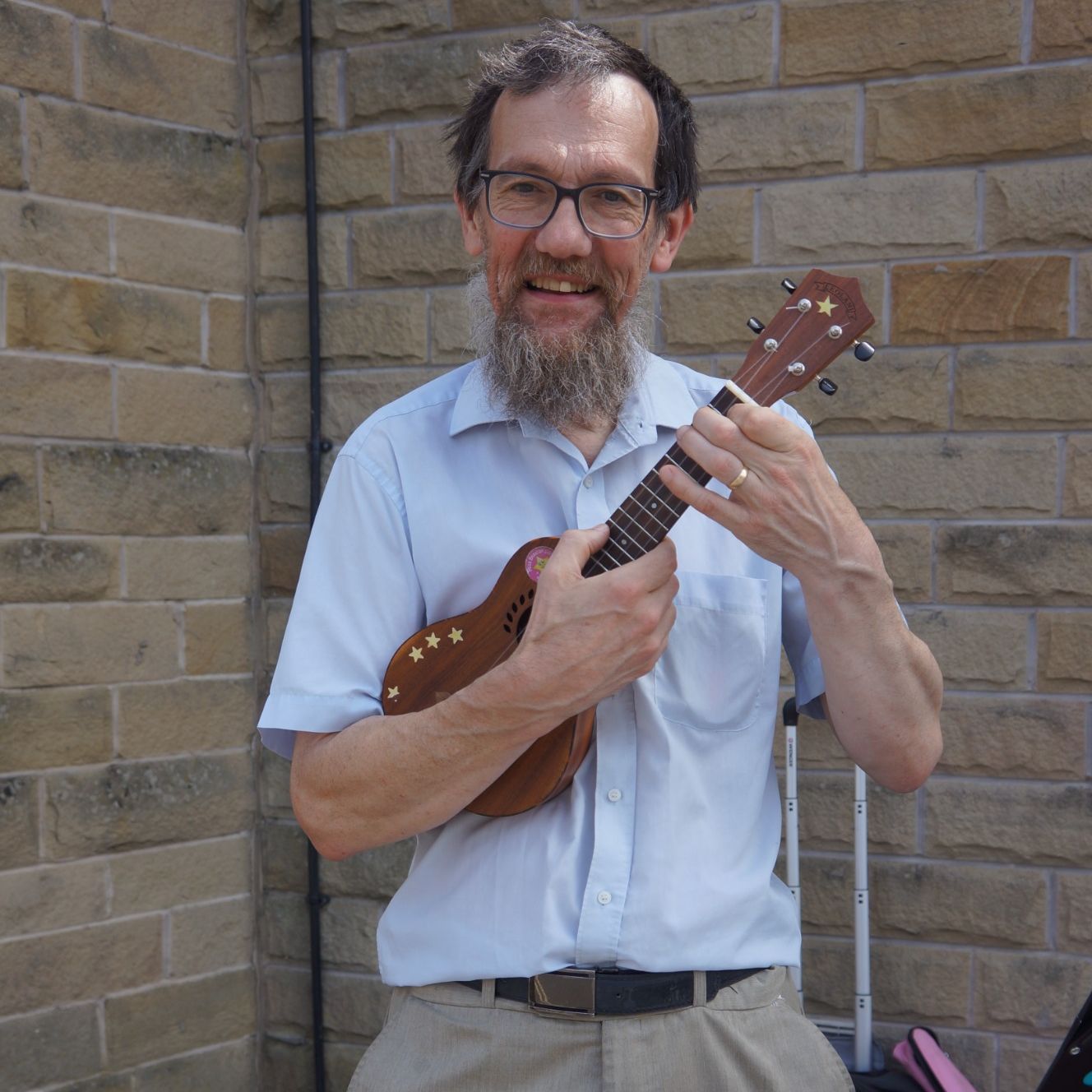 Eric, wearing red sparkly hat, in campsite, holding up his hand, with T shirt with the logo 'Never underestimate an old man with a ukulele'